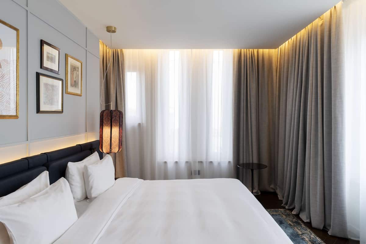 A hotel room with a large bed, a headboard with a light fixture, a small table, and a window with curtains.
