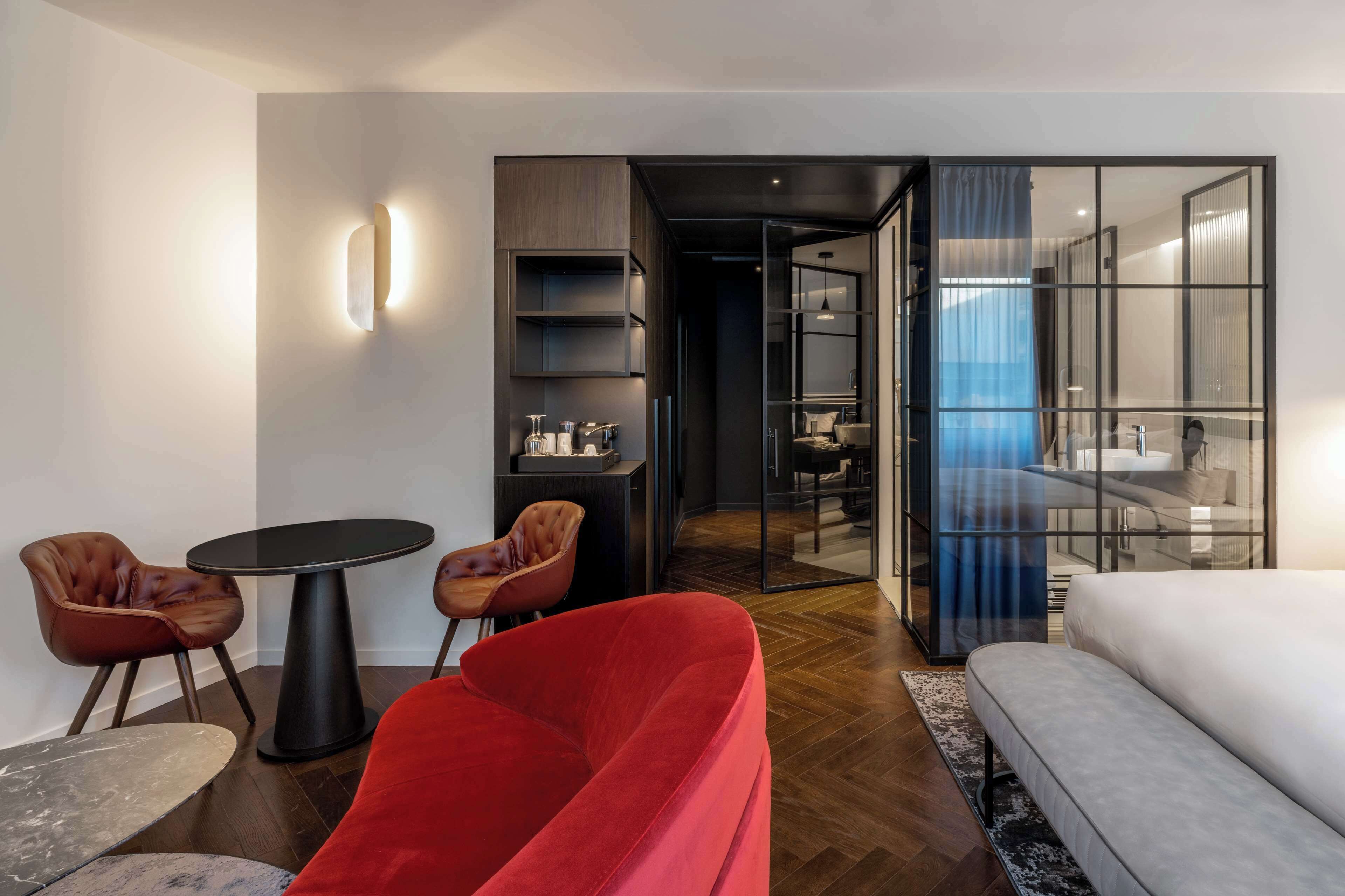 Una camera d'hotel moderna con un divano rosso, un letto bianco, un tavolino rotondo con due sedie e un pannello di vetro che separa la stanza dal bagno.