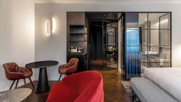 Una camera d'hotel moderna con un divano rosso, un letto bianco, un tavolino rotondo con due sedie e un pannello di vetro che separa la stanza dal bagno.