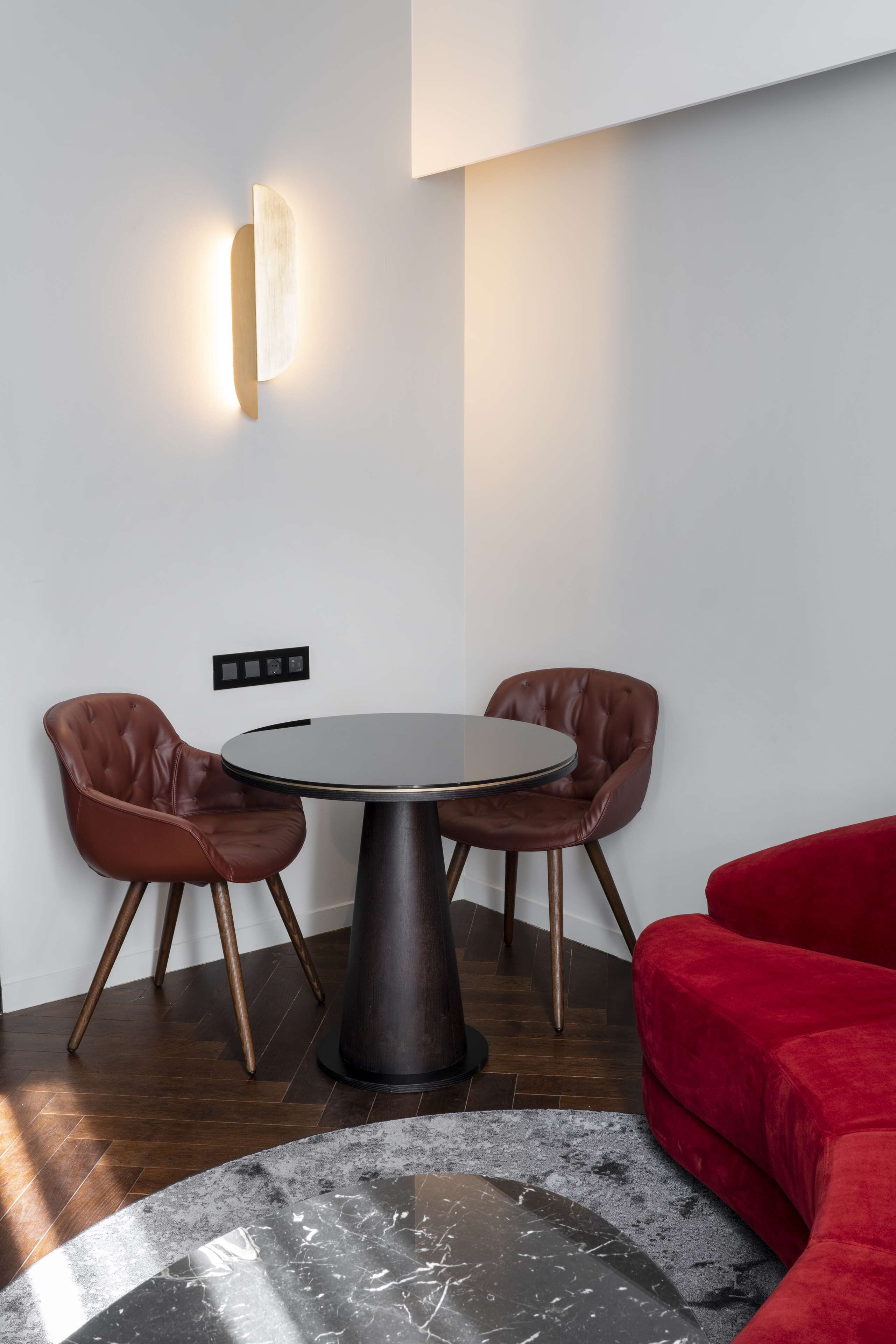 A modern dining area with a round table, two leather chairs, a red sofa, and a wall-mounted light fixture.