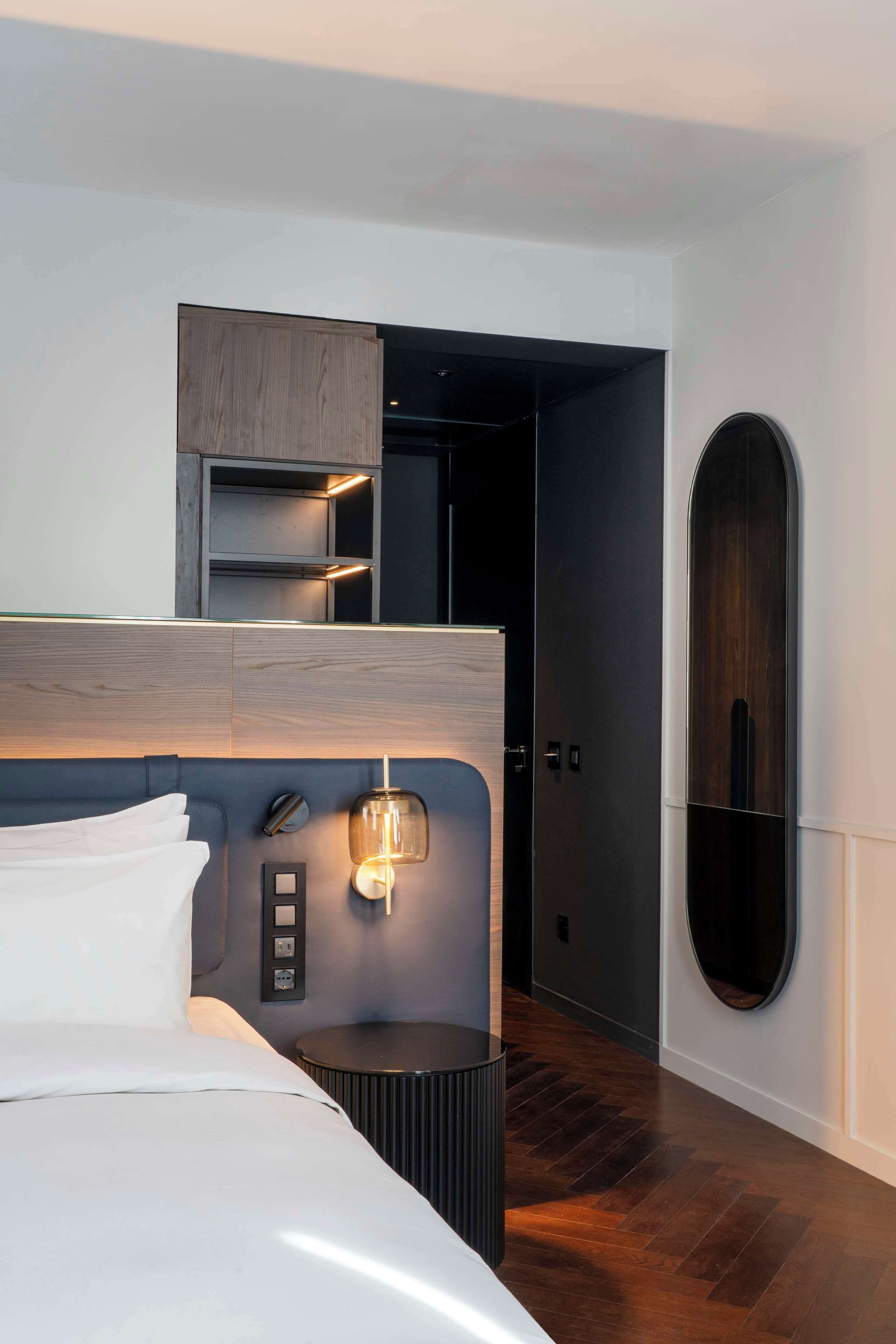 A modern hotel room with a large bed, a bedside table, a wall-mounted shelf, and a door leading to another room.
