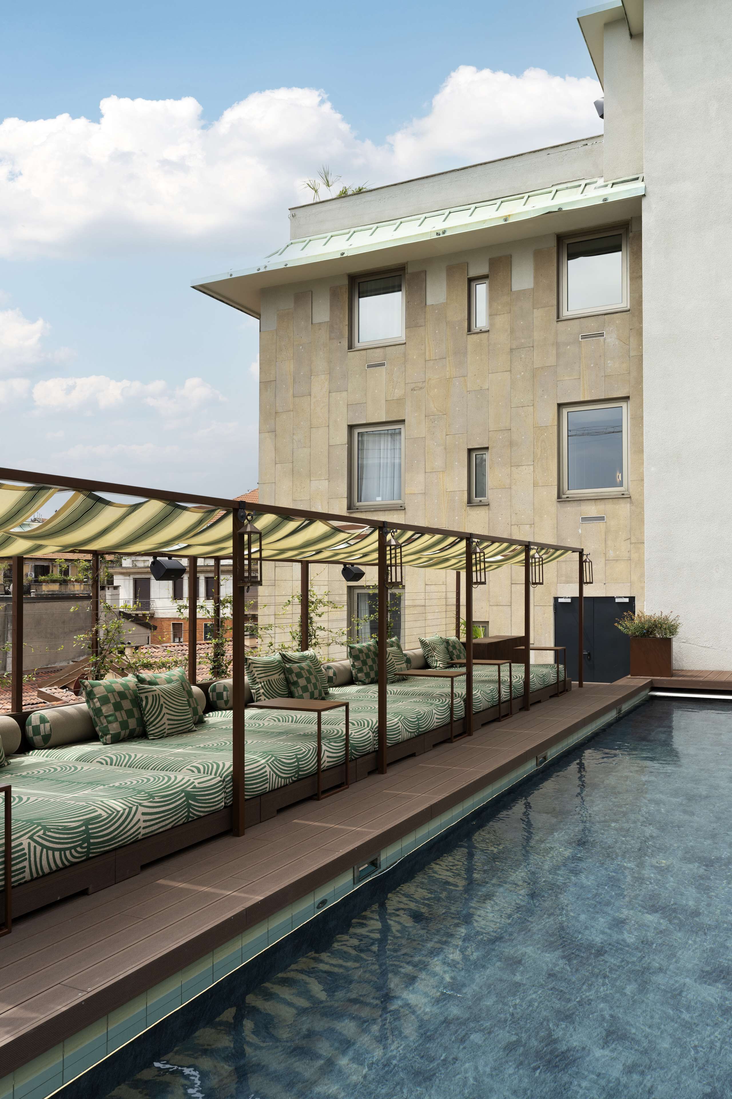 Un espace aménagé au bord de la piscine, avec un coin salon couvert, une piscine à eau claire et un bâtiment doté de grandes fenêtres en arrière-plan.
