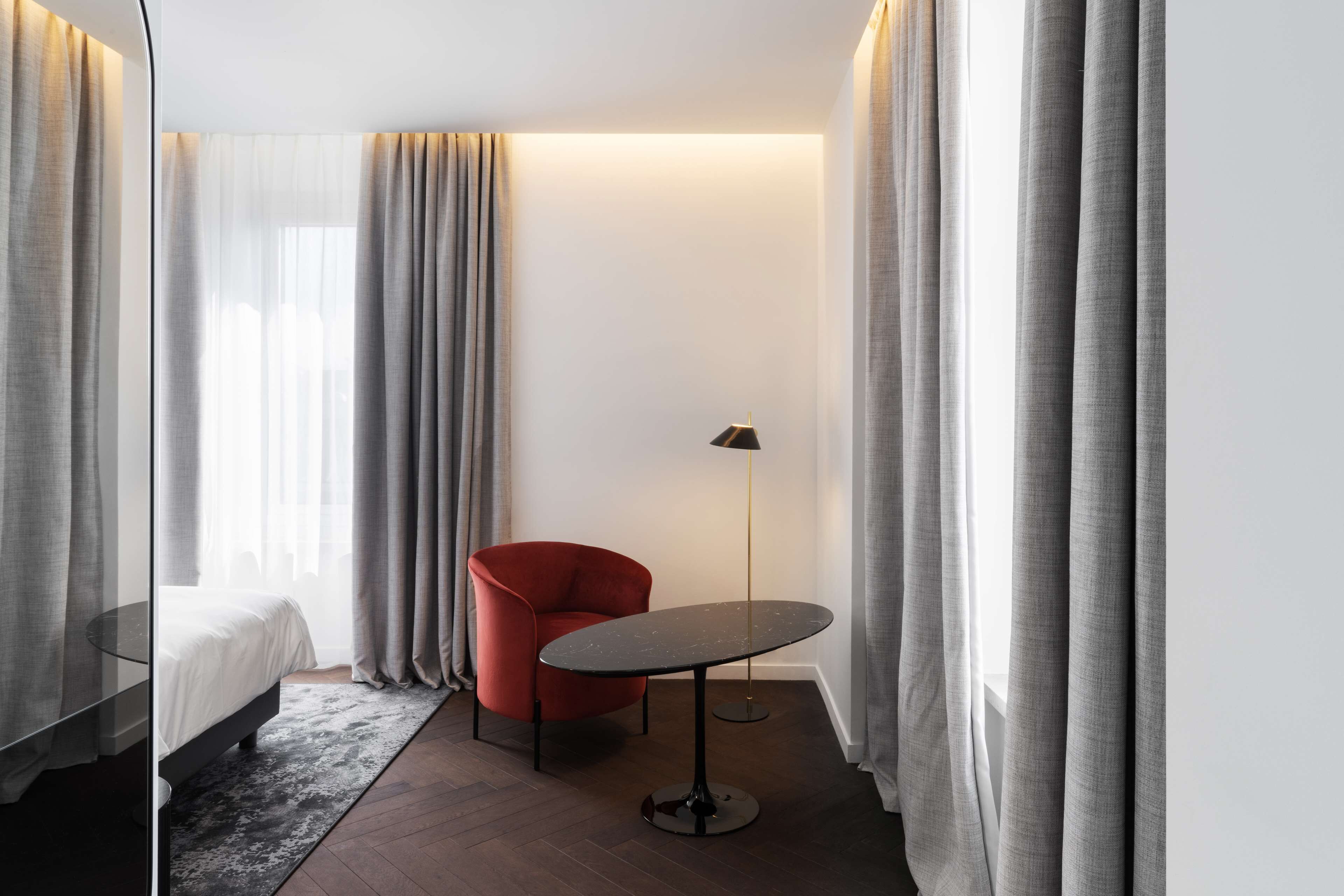 A hotel room with a bed, a red chair, a small round table, and a floor lamp.