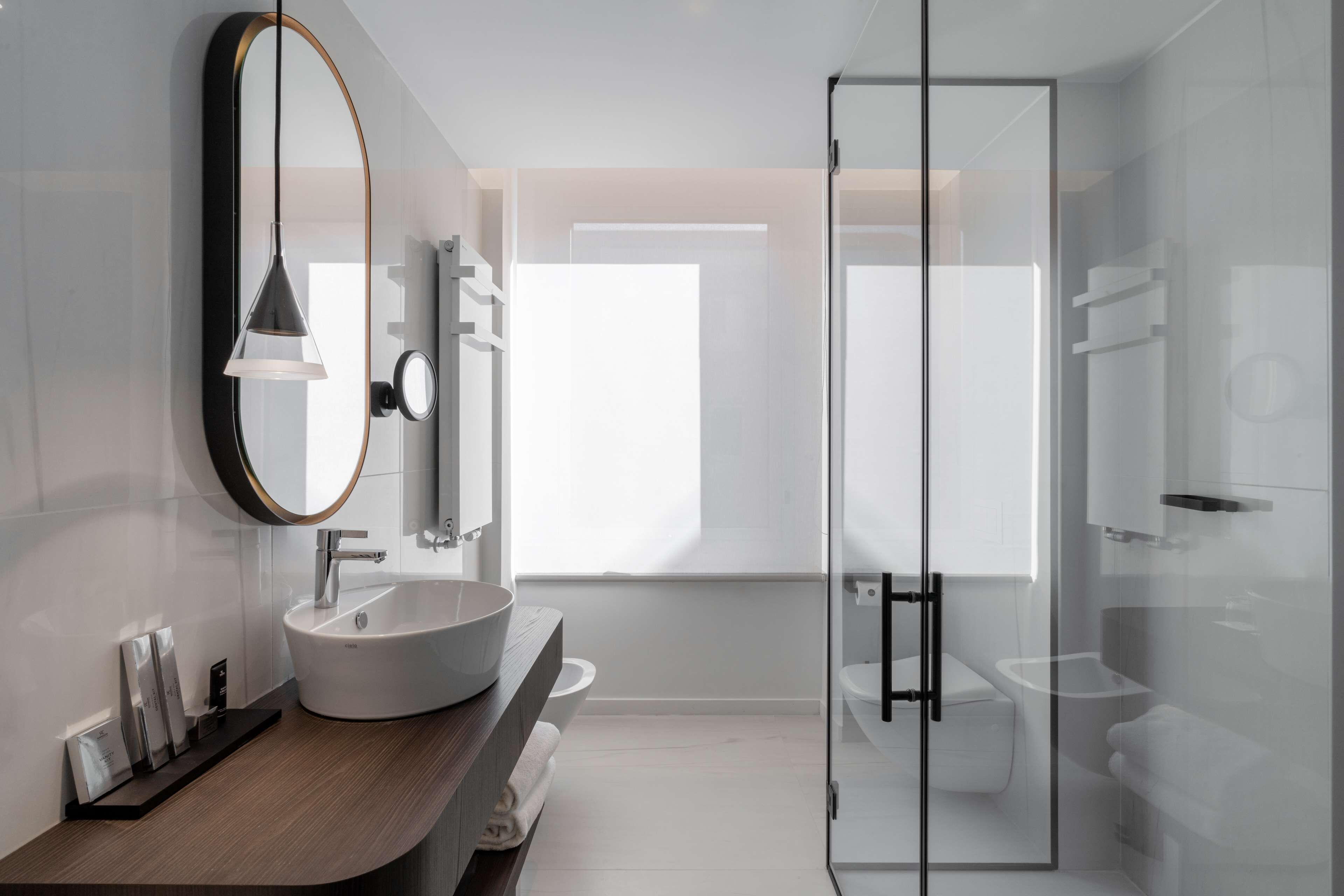 A modern bathroom with a large oval mirror, a white sink, and a glass-enclosed shower.