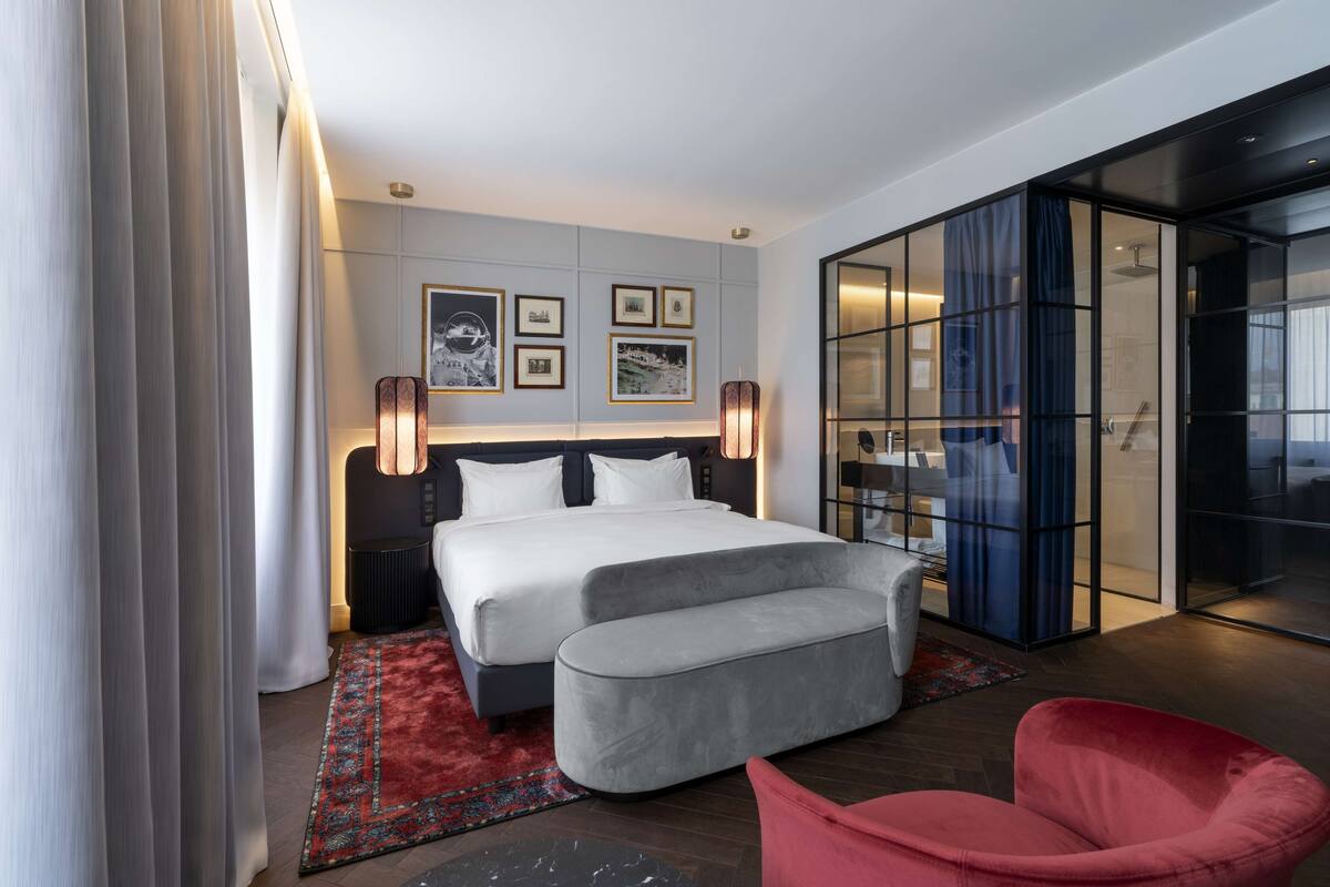 A modern hotel room with a large bed, a red armchair, and a glass partition.