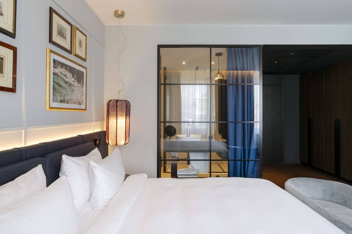 A modern hotel room with a large bed, a bedside table, a mirror, and a view of a bathroom through a glass partition.