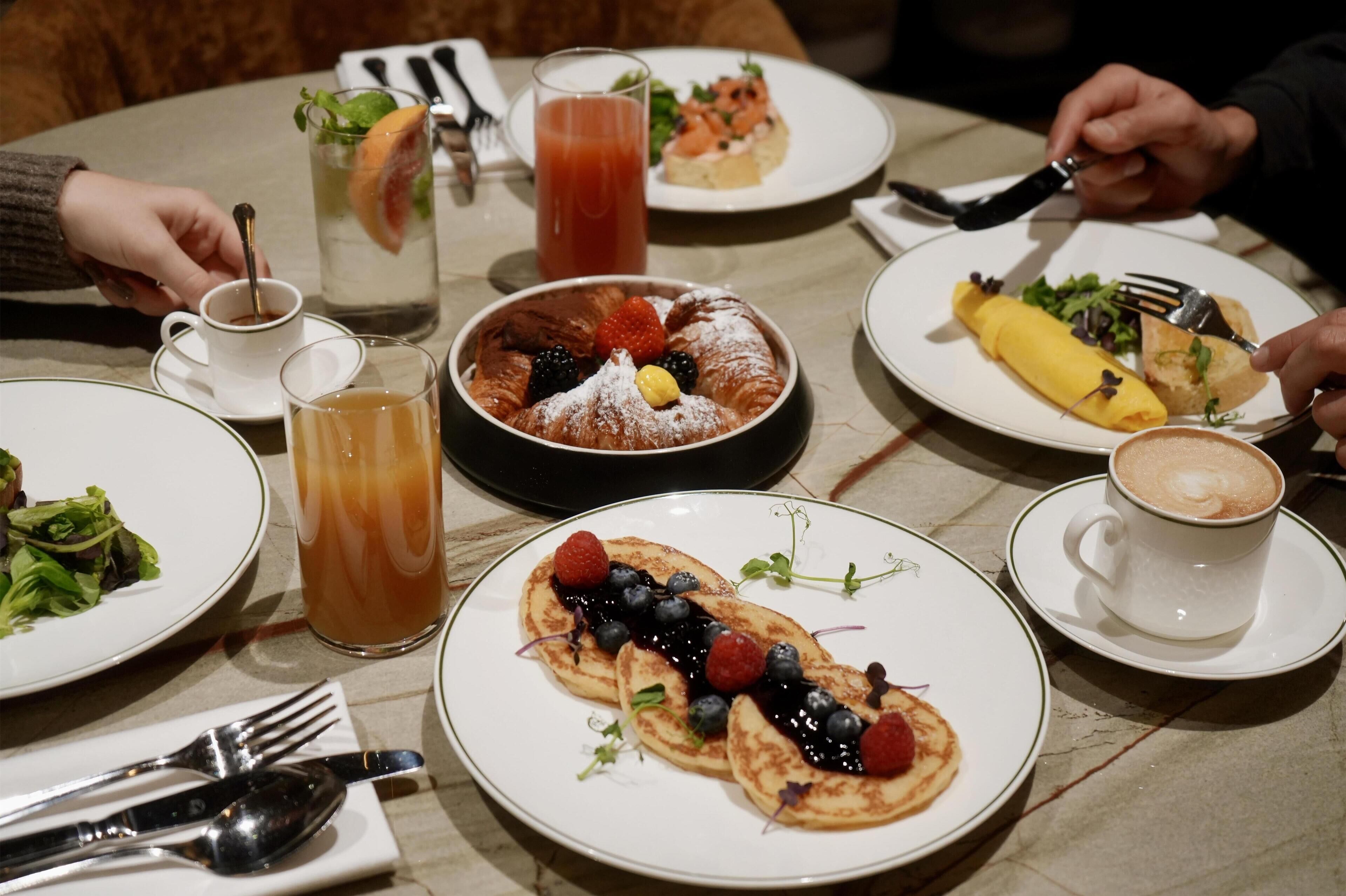 Ein Esstisch mit verschiedenen Speisen, darunter Pfannkuchen mit Beeren, Obstsalat und ein Glas Eistee.