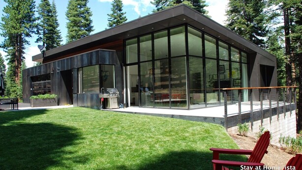 Une maison moderne avec de grandes fenêtres en verre, un barbecue et une chaise rouge au premier plan.