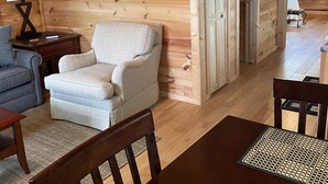A cozy cabin interior with a plaid armchair, a wooden dining table, and a lamp on a nightstand.