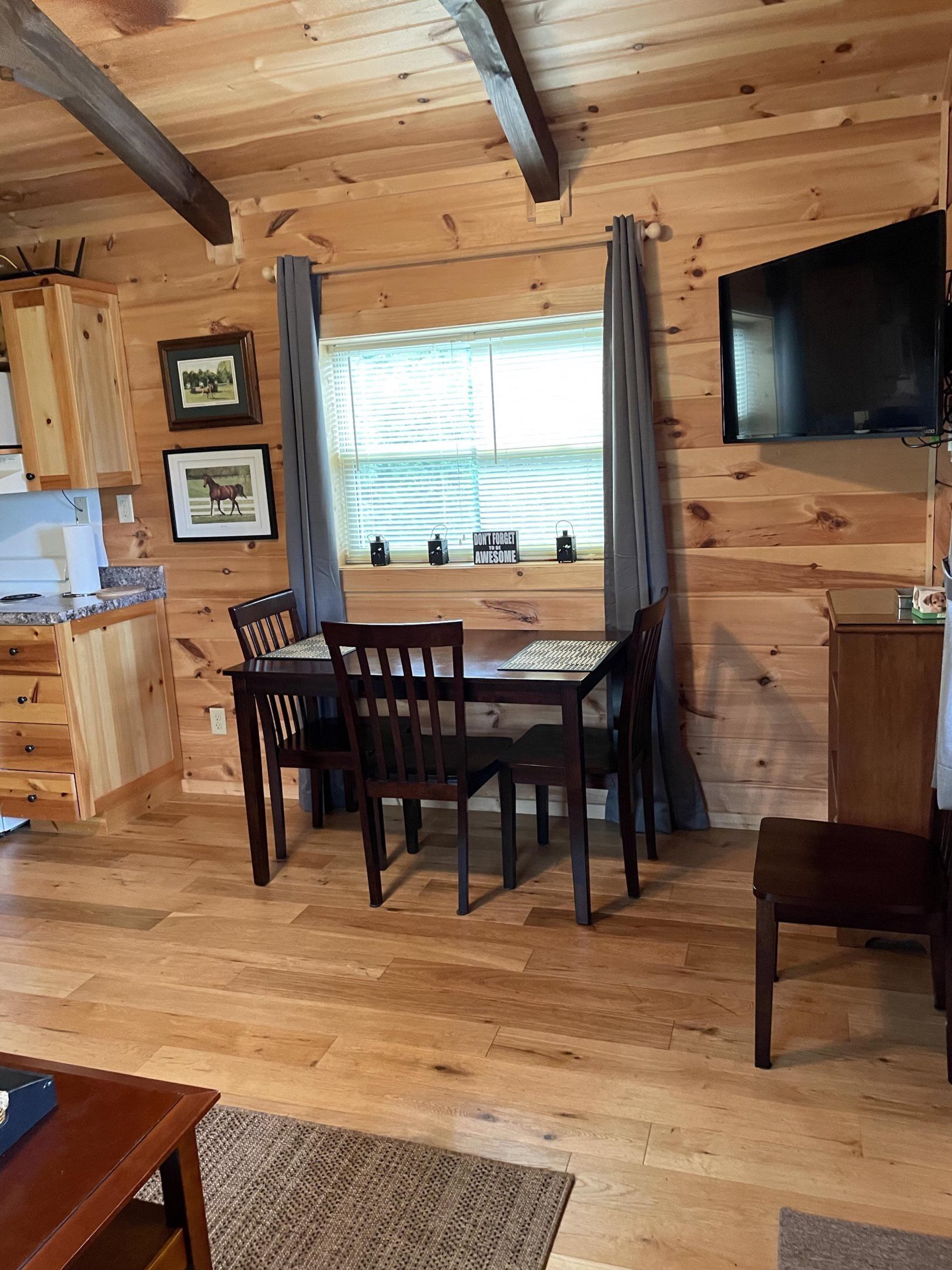 A wooden cabin interior with a dining table, chairs, a kitchen area, and a television mounted on the wall.