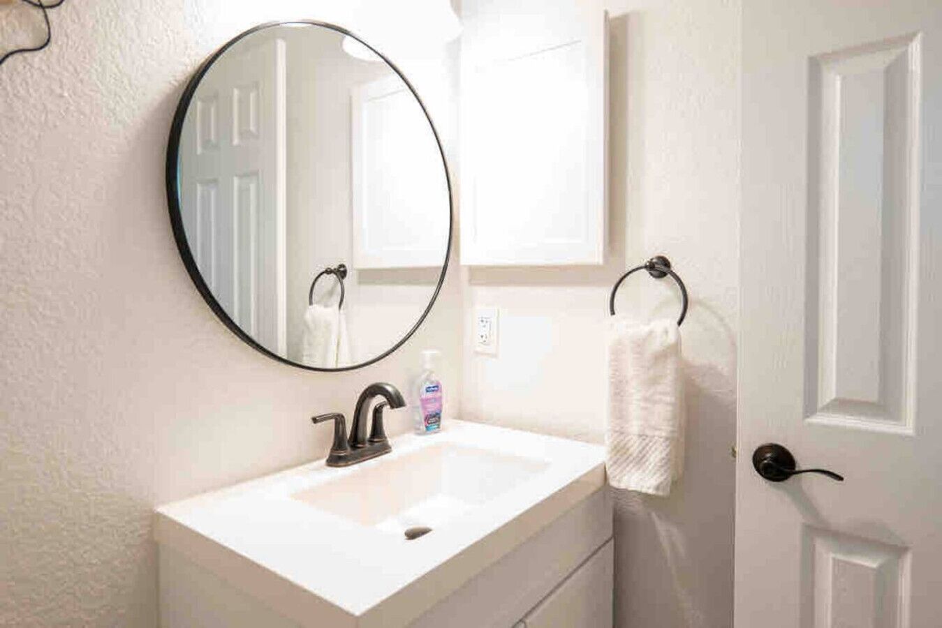 A bathroom with a round mirror, a white sink, a black faucet, a towel rack, and a door.
