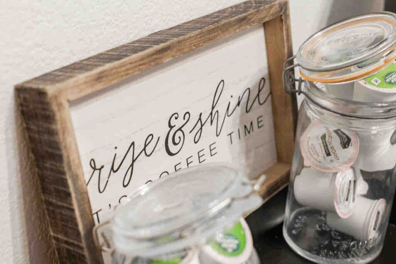 Uma placa de madeira com a inscrição 'Rise & Shine' e um pote de café com etiqueta verde.