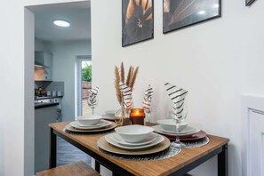 Ein Esszimmer mit Tisch, Geschirr, Gläsern und Dekorationselementen, mit Blick aus einer modernen Küche.