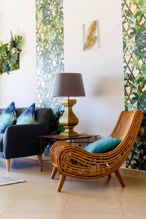 A living room with a black sofa, a wicker chair, a table with a lamp, and a wall with a leafy pattern. - Boavista Golf Resort Apartment (Lagos)