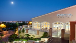 Un hotel llamado 'Essential Fitness & Spa' con una entrada bien iluminada, zona de estar al aire libre y vistas a la ciudad de fondo.