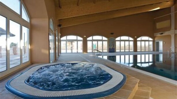 Una amplia piscina interior con un jacuzzi en el centro, rodeada de grandes ventanales y un techo de madera.