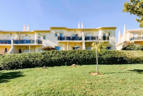 A row of yellow buildings with balconies, a grassy area in front, and a hedge.