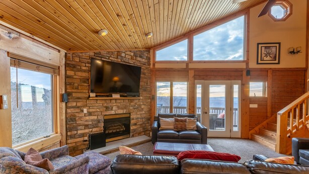 A spacious living area with a stone fireplace, leather sofas, a flat-screen TV, and large windows offering a view of the outdoors.