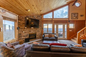A spacious living area with a stone fireplace, leather sofas, a flat-screen TV, and large windows offering a view of the outdoors.