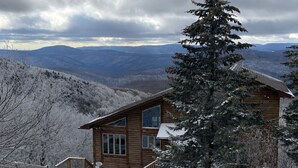 A wooden cabin with a staircase, surrounded by snow-covered trees and mountains.