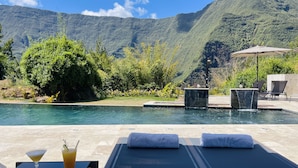 Un área junto a la piscina con sillones reclinables, una mesa con bebidas y vistas a la montaña.