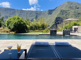 Un área junto a la piscina con sillones reclinables, una mesa con bebidas y vistas a la montaña.