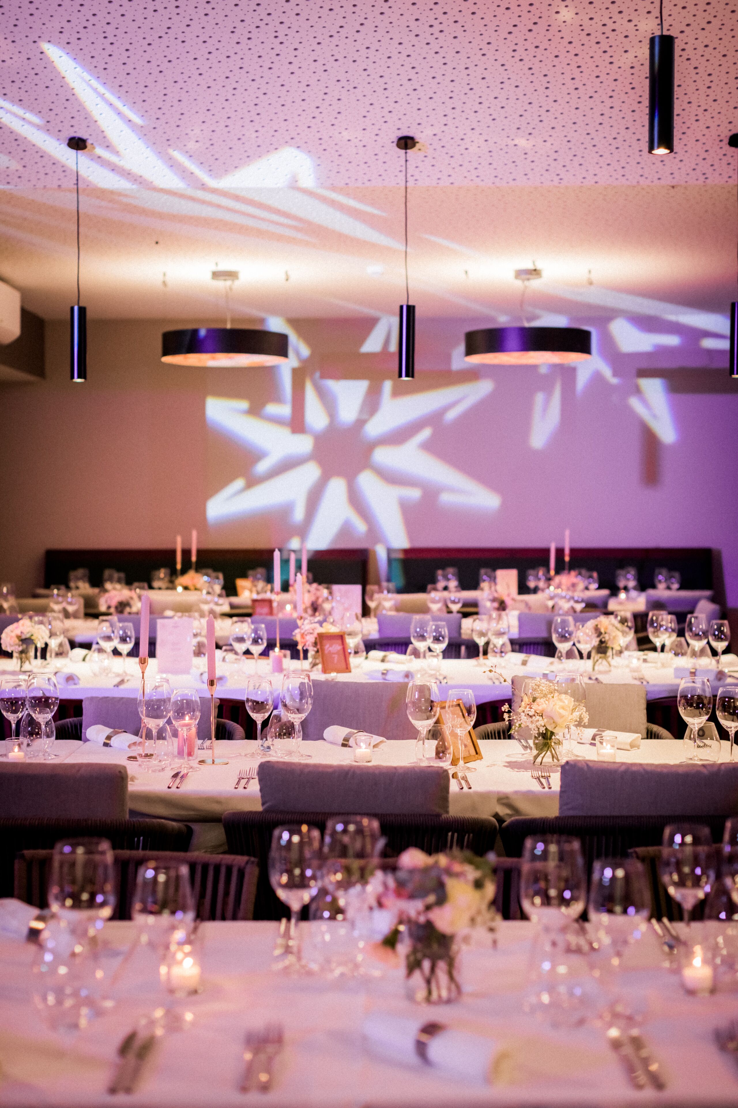A banquet hall with tables set for a formal event, featuring overhead lighting and a projection screen in the background.