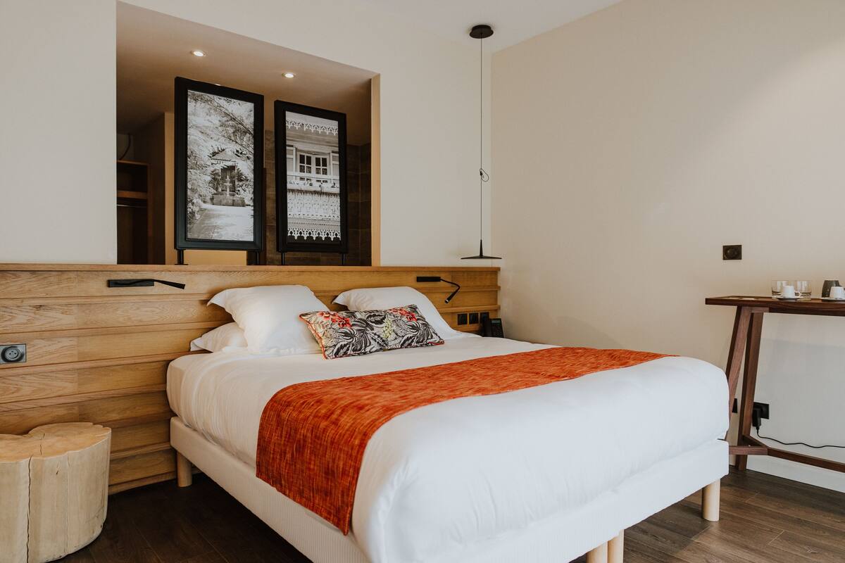 A modern hotel room with a wooden headboard, a large bed with white bedding and an orange throw, a bedside table with a lamp, and two framed pictures above the bed.