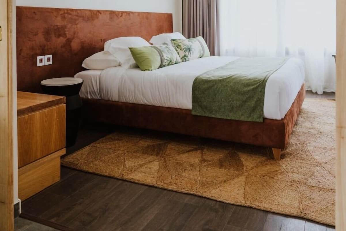 A modern hotel room with a wooden headboard, a large bed with white bedding and green accents, a round bedside table, and a wooden nightstand.