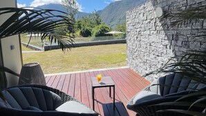 Un área de estar al aire libre techada con sillas de mimbre y una pequeña mesa, con vista a un muro de piedra y un patio cubierto de césped, con montañas al fondo.