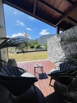 Un área de estar al aire libre techada con sillas de mimbre y una pequeña mesa, con vista a un muro de piedra y un patio cubierto de césped, con montañas al fondo.