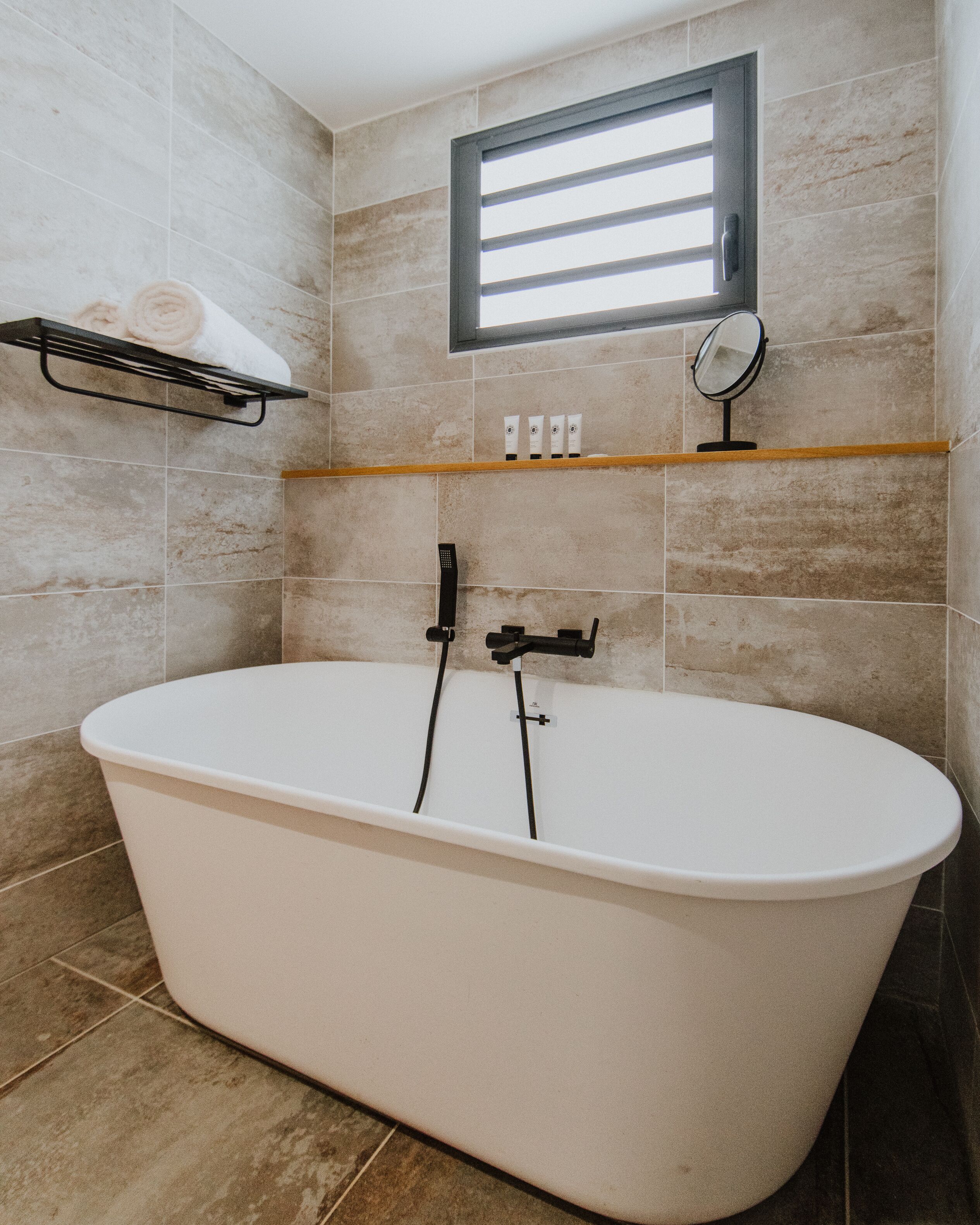 Un baño moderno con una bañera independiente, un perchero con una toalla enrollada, un estante con artículos de aseo y una ventana con persianas.