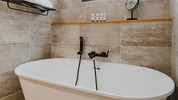 Un baño moderno con una bañera independiente, un perchero con una toalla enrollada, un estante con artículos de aseo y una ventana con persianas.