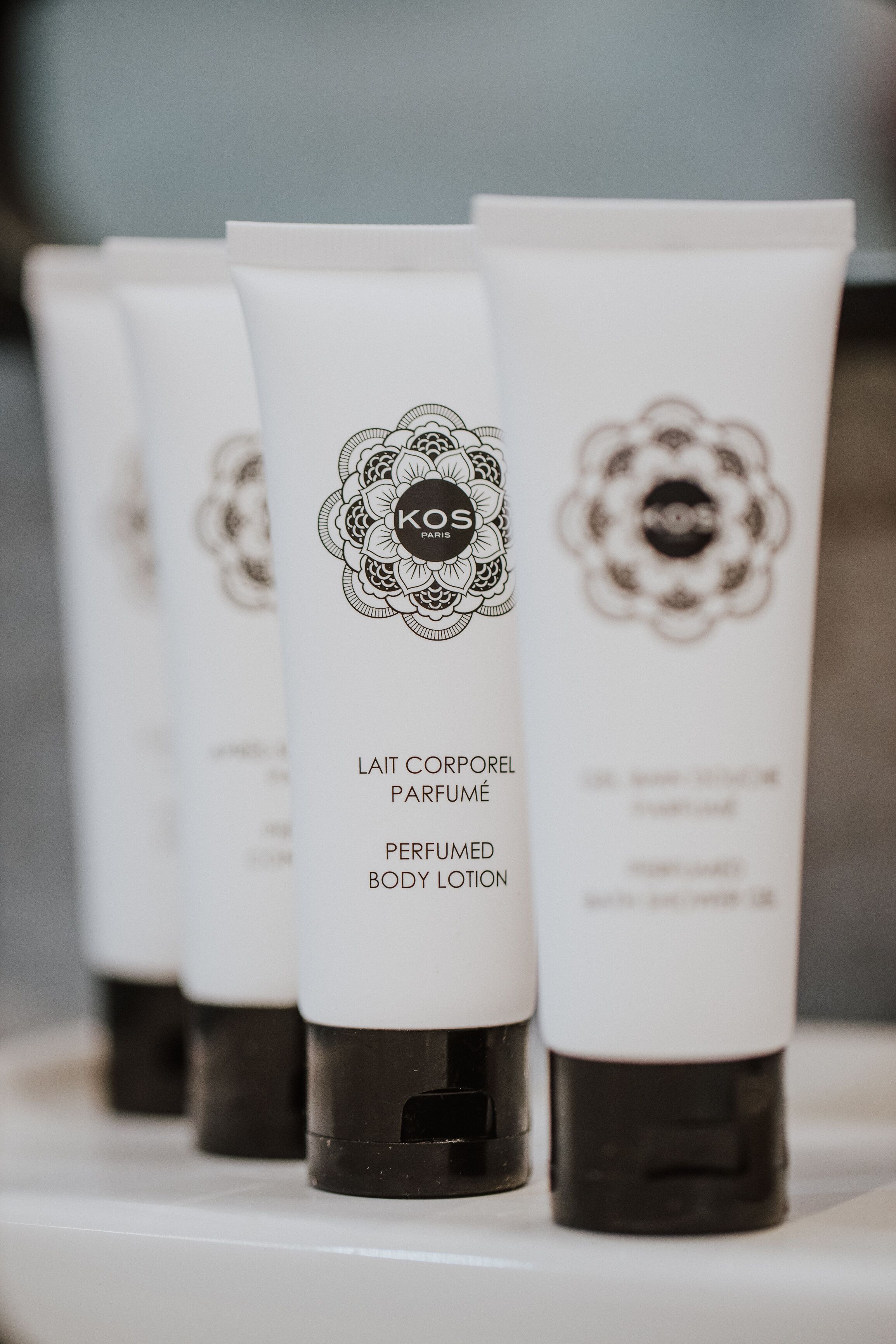 A row of white lotion tubes with black caps, labeled "KOS PARIS" and "LAIT CORPOREL PARFUMÉ PERFUMED BODY LOTION".