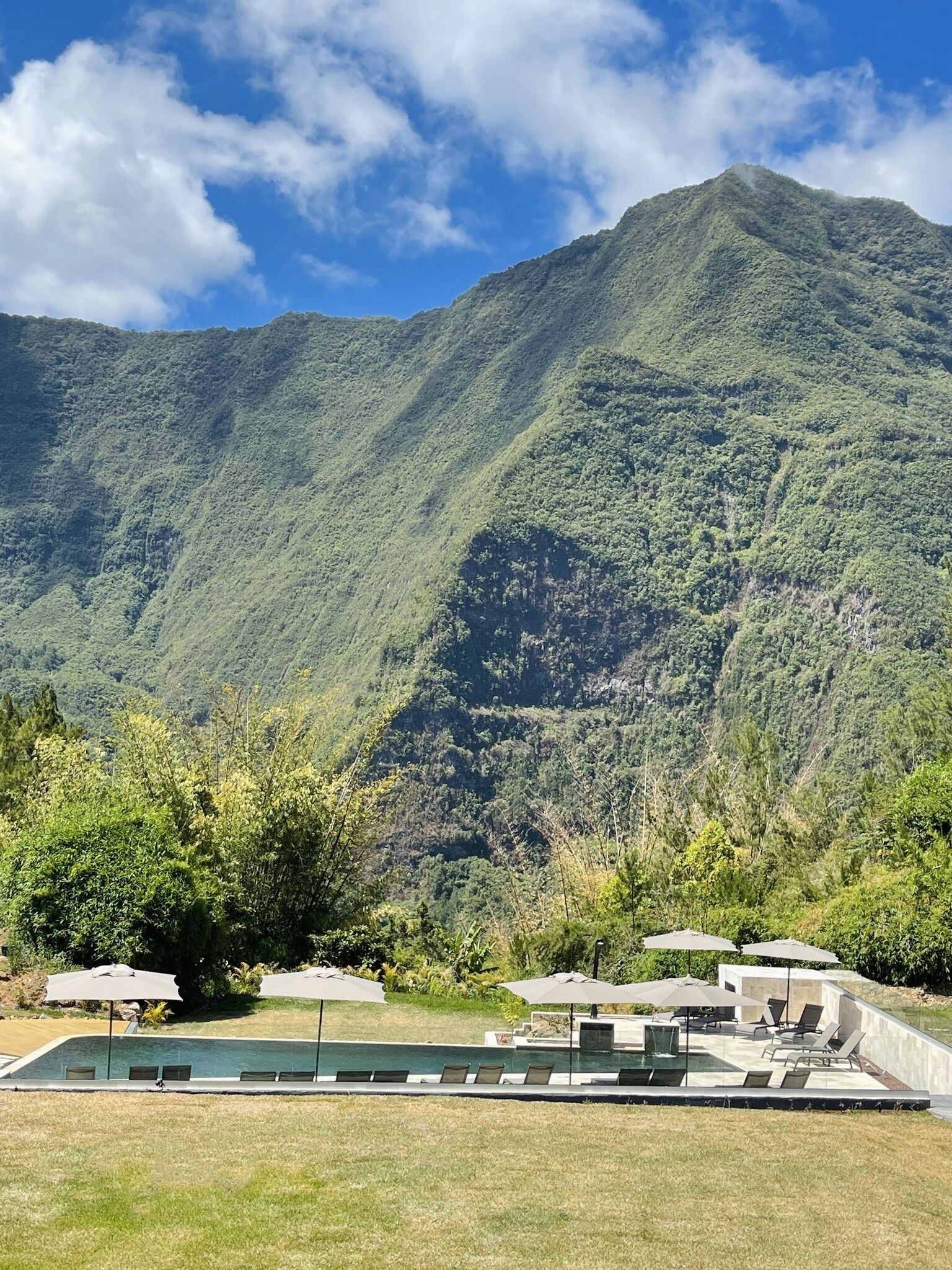 Zona de piscina con sillones de descanso y sombrillas, rodeada de abundante vegetación y montañas al fondo.