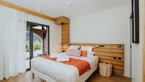 Un dormitorio moderno con cabecera de madera, una cama grande con ropa de cama blanca y detalles naranja, un taburete de madera pequeño y vistas a zonas verdes a través de una puerta corrediza de vidrio.