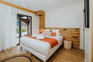 Un dormitorio moderno con cabecera de madera, una cama grande con ropa de cama blanca y detalles naranja, un taburete de madera pequeño y vistas a zonas verdes a través de una puerta corrediza de vidrio.