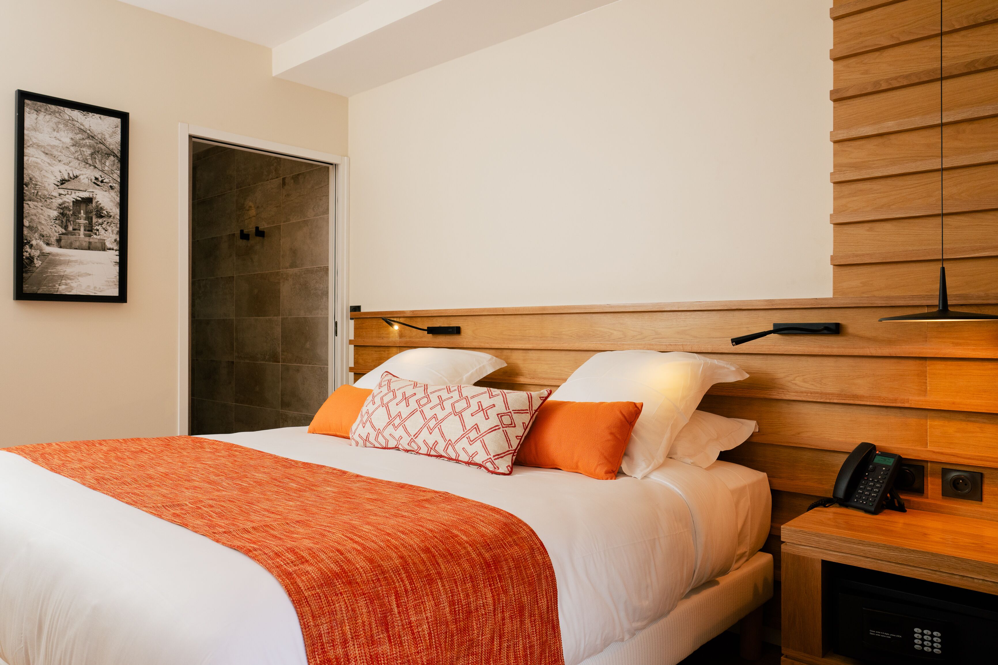 Habitación de hotel con cama, mesita de noche con teléfono, puerta y lámpara fijada a la pared.