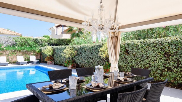 Un área de comedor al aire libre con mobiliario de mimbre, piscina y un seto de fondo.