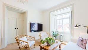 A bright, white room with a large window, a flat-screen TV, wicker chairs, a wooden table with a plant, and a bed with a floral pillow. - Casa Omlandia With Pool - Lagos Old Town (Lagos)