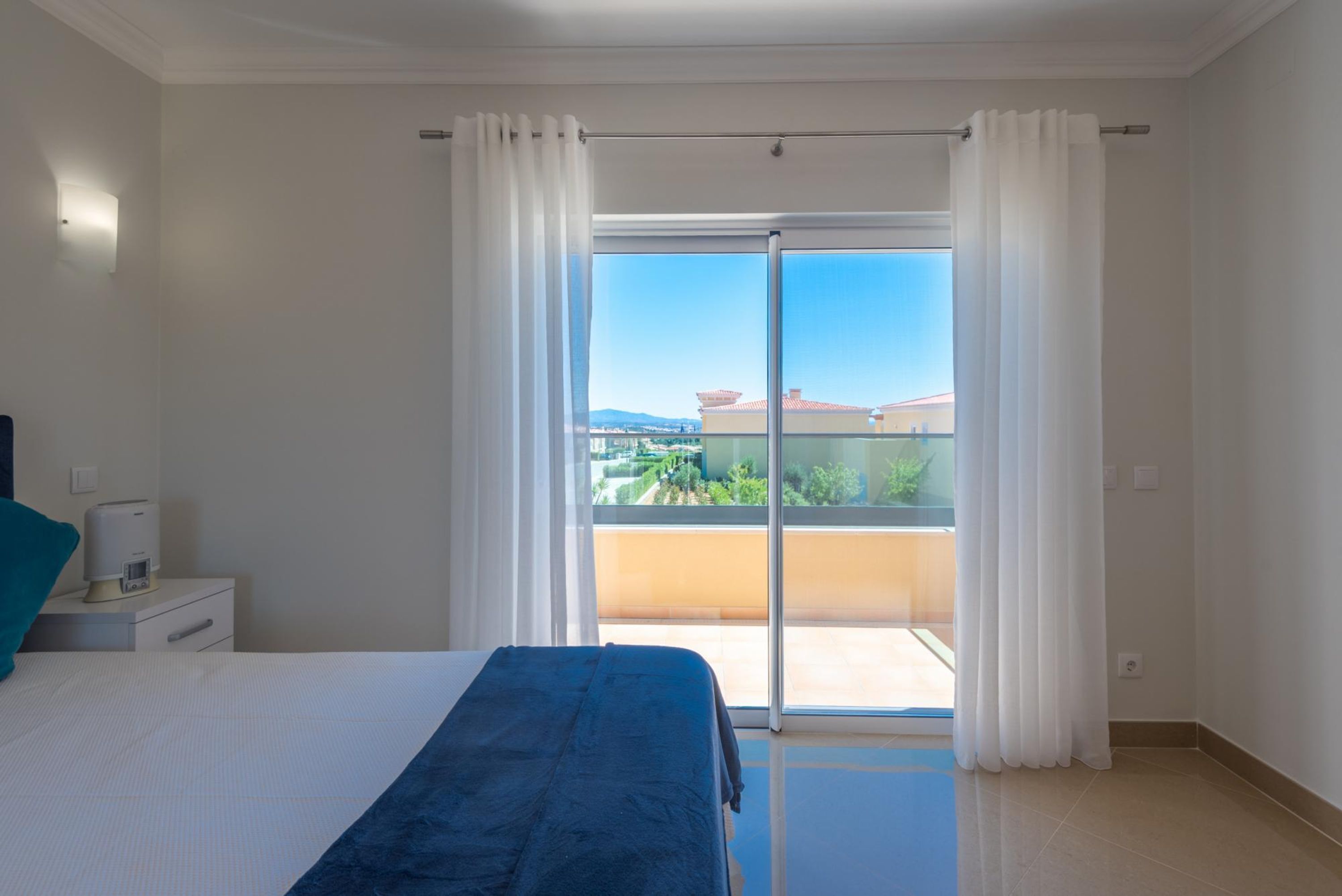 Une chambre à coucher avec un lit, une table de chevet, une fenêtre avec des rideaux et une vue sur un balcon agrémenté de verdure et entouré d’immeubles.
