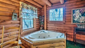 Ein Badezimmer einer Holzhütte mit einer großen Badewanne, einem Holzstuhl, einem Fenster mit Blumenvorhang und einem gerahmten Gemälde.