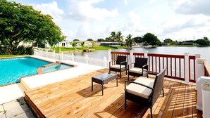 A wooden deck with a pool, outdoor furniture, and a view of a lake and distant buildings. - Stunning Lake-Front Villa with Pool & Spa (Pembroke Pines)