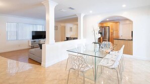 A modern living room with a glass dining table, white chairs, and a TV. - Stunning Lake-Front Villa with Pool & Spa (Pembroke Pines)