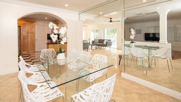 A modern dining area with a glass table and white chairs, a kitchen in the background, and a large mirror reflecting the space. - Stunning Lake-Front Villa with Pool & Spa (Pembroke Pines)
