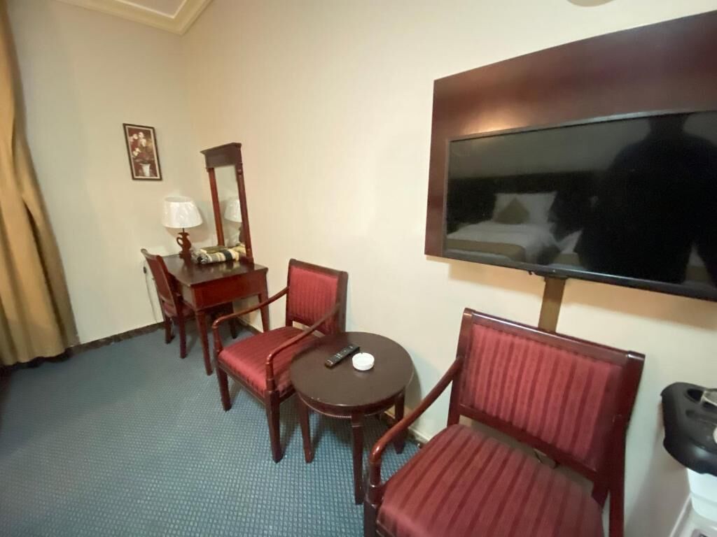 A hotel room with a bed, a TV mounted on the wall, a desk with a chair, a small table with a chair, and a lamp.
