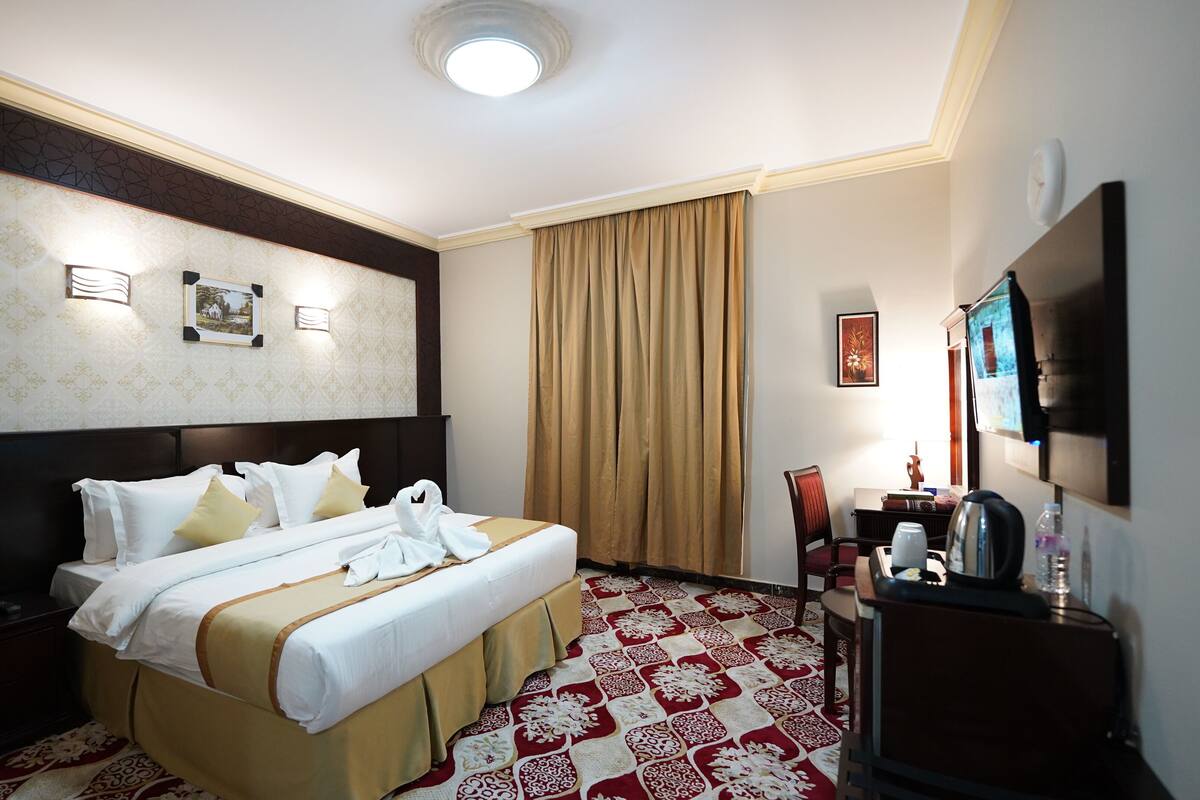A hotel room with a large bed, a desk with a chair, a flat-screen TV, a telephone, and a decorative wall panel.