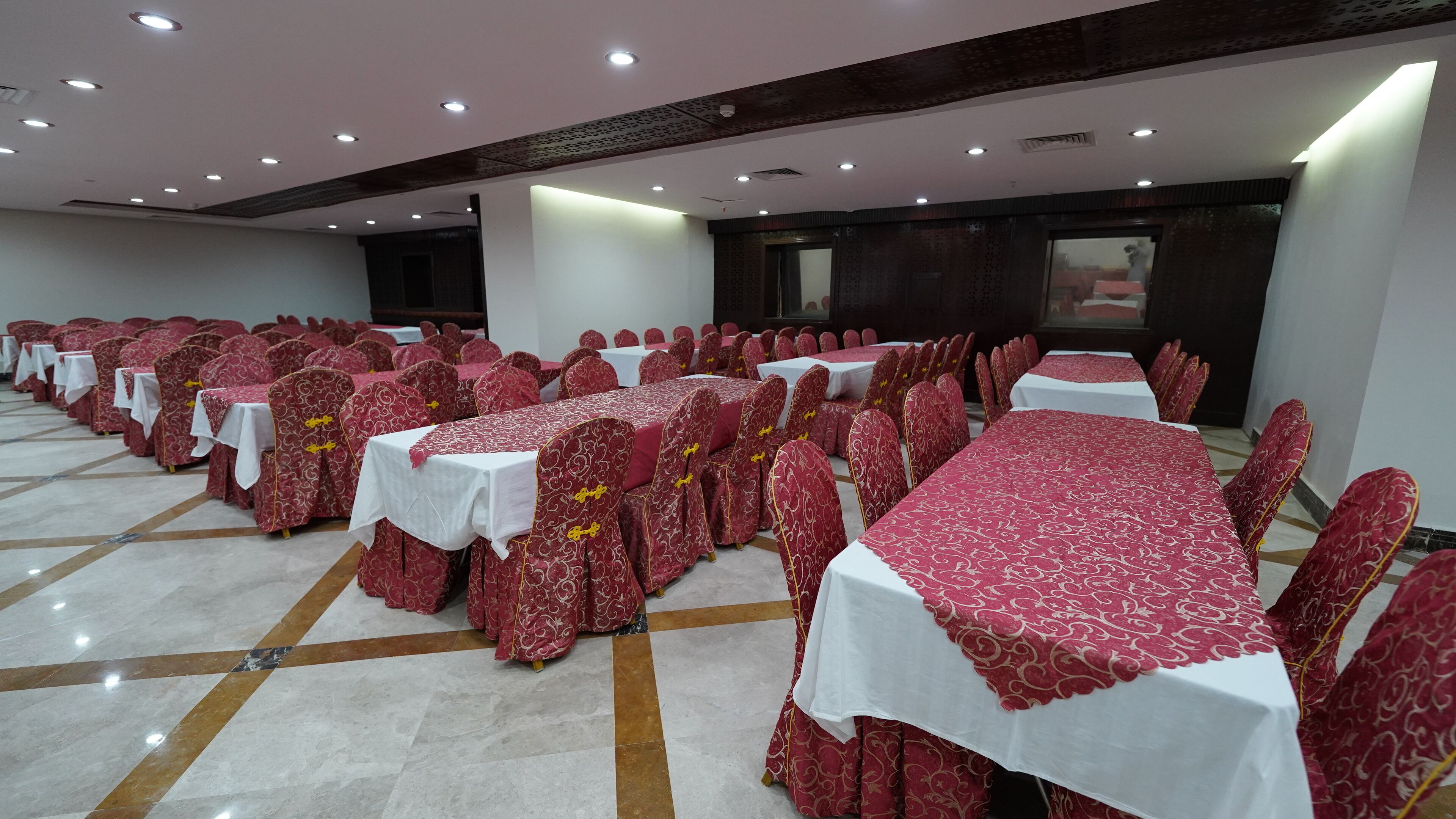 A banquet hall with rows of tables and chairs, each set with a white tablecloth and red patterned chair covers.