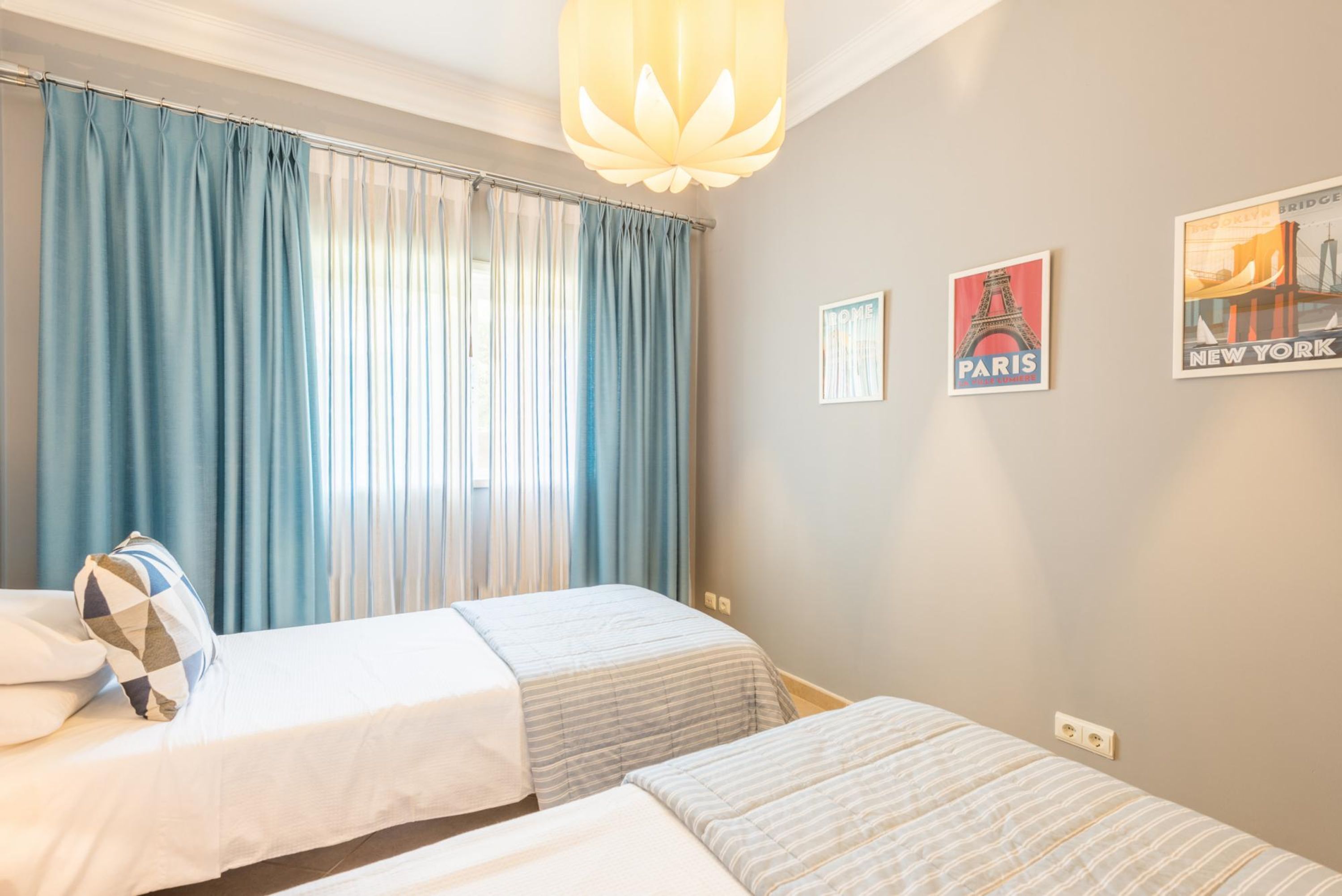 A hotel room with two beds, a window with blue curtains, a hanging light fixture, and two posters on the wall.
