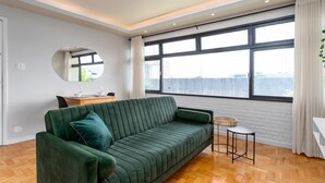 A modern living room with a green sofa, a round mirror, a small side table, and a large window with curtains.